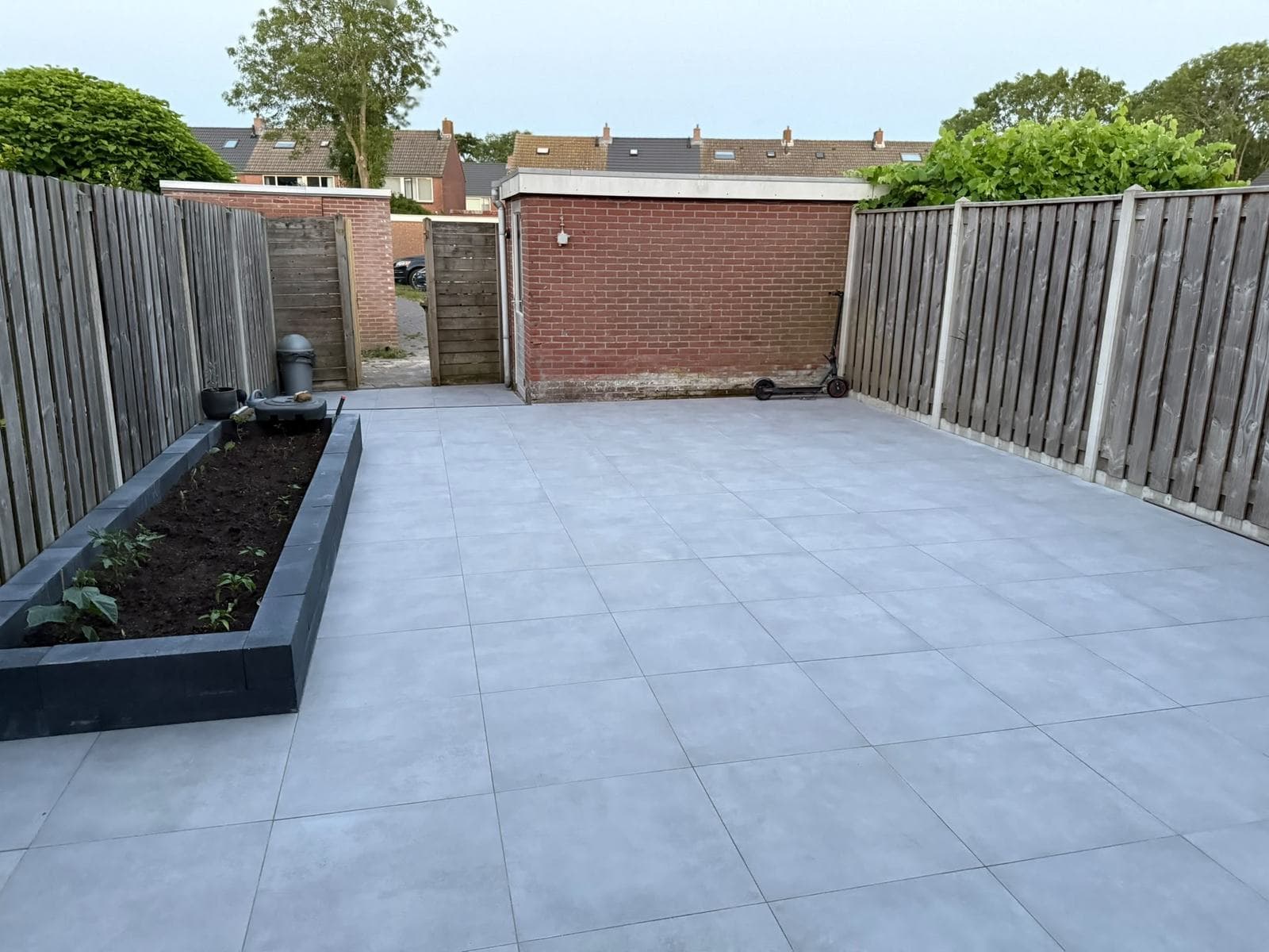 Dezelfde achtertuin na renovatie: strakke tegelvloer, nieuwe schutting en een gemetselde plantenbak